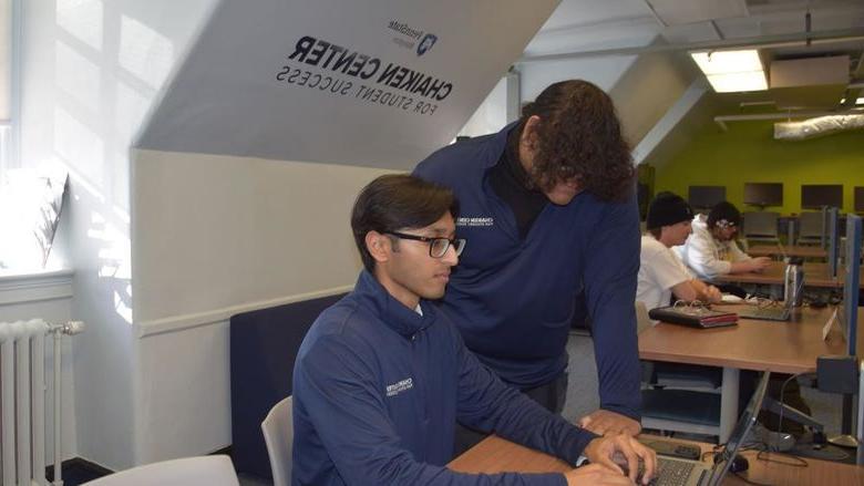 Person standing and person sitting and looking at a laptop